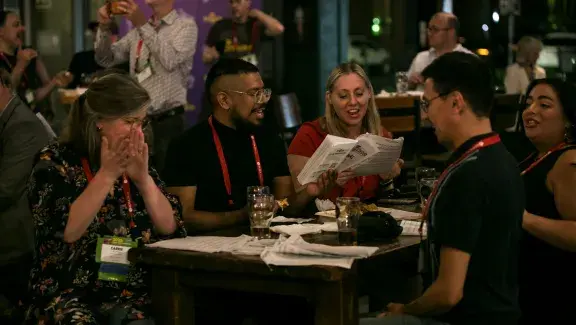 group of people sitting at a table and singing joyfully with empty beer cups by them. One of them holds sheet music up as he sings