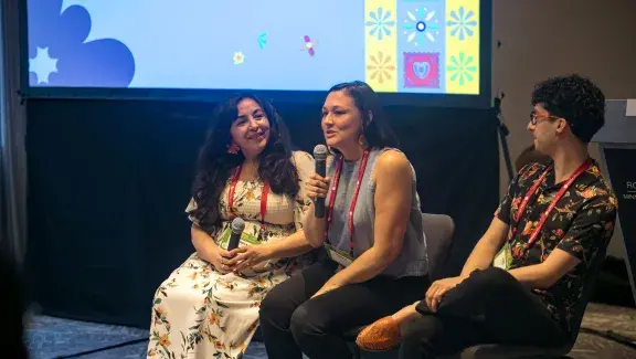 Three people sitting in a row as a panel. The person in the middle has a microphone and is talking. The two on either side are turned towards her listening as she speaks